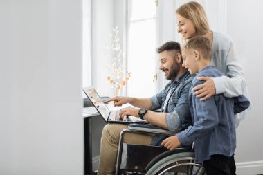 Mutlu anne gülümseyen oğlunu kucaklıyor ve evinde tekerlekli sandalyede oturan sakallı kocasını tatmin ediyor. Dost canlısı aile, pencerenin yanında dizüstü bilgisayar kullanıyor..