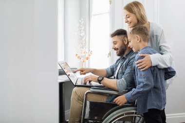 Mutlu anne gülümseyen oğlunu kucaklıyor ve evinde tekerlekli sandalyede oturan sakallı kocasını tatmin ediyor. Dost canlısı aile, pencerenin yanında dizüstü bilgisayar kullanıyor..