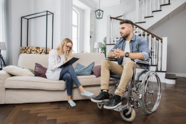 Hasta ile rahat bir kanepede otururken, panoda rehabilitasyon programına odaklanmış bir fizyoterapist. Tekerlekli sandalyedeki engelli adam zaman çizelgesine katılıyor..