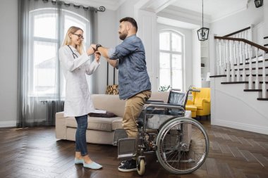 Pozitif genç terapist, rahat evinde tekerlekli sandalyeden kalkması için yakışıklı sakallı adama yardım ediyor. Tatmin olmuş bir adam yürümeye çalışırken bacaklarındaki kasları hissetmeye başlıyor..
