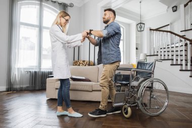 Gözlüklü ve beyaz laboratuvar önlüklü sağlık görevlisi engelli erkek hastaların modern dairede tekerlekli sandalyeden kalkmalarına yardım ediyor. Başarılı rehabilitasyon kavramı.