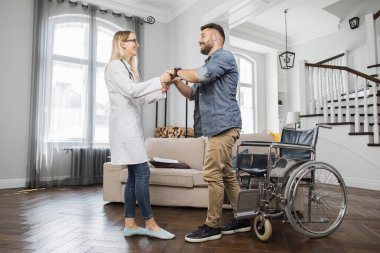 Profesyonel kadın terapist sakallı adamın tekerlekli sandalyeden kalkıp eve yürümesine yardım ediyor. Engelli neşeli erkek kazadan sonra iyileşiyor..