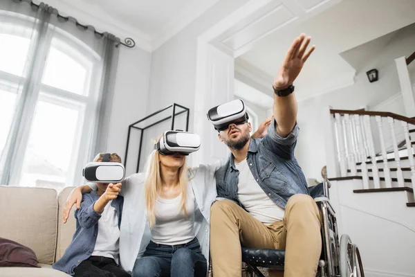 Disabled caucasian man with wife and son wearing virtual reality glasses and playing video game in living room. Lovely family enjoying imagination activity together.