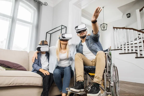 Happy caucasian family with disabled father in wheelchair playing games together using VR headsets. Modern technology for entertainment at home.