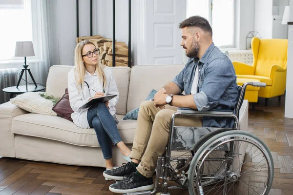 Female healthcare worker with clipboard sitting on comfortable sofa and examining caucasian man in wheelchair at home. Nurse discussing with patient rehabilitation course.