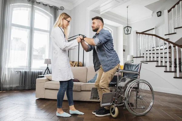 Caring health care worker in eyeglasses and white lab coat helping disabled male patient to get up from wheelchair at modern apartment. Concept of successful rehabilitation.