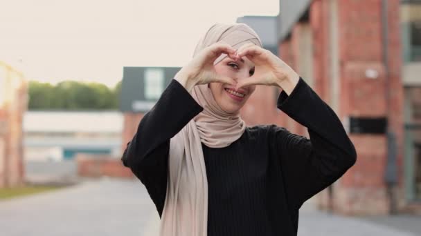 ヒジャーブのアラブ系イスラム教徒の女性と都市の背景にポーズをとる黒い服を笑顔 宗教的なイスラームライフスタイルの概念 手でハートの形を見せる ハートの形をしたサイン — ストック動画