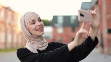 Gülümseyen mutlu Müslüman kadının dijital akıllı telefon kullanarak rahatlamasının portresi. Genç Müslüman kız şehirdeki sosyal medyada çevrimiçi video görüşmesi sırasında ekranda konuşma yapıyor..