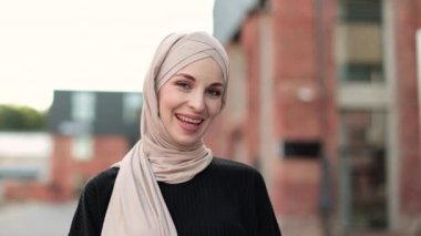 Portrait of young muslim woman wearing hijab head scarf in city while looking at camera. Closeup face of cheerful woman covered with headscarf smiling outdoor. Casual islamic girl in town.