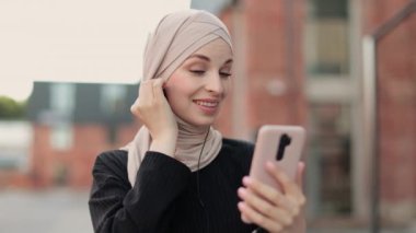 Genç Müslüman kadın şehir caddesinde yürürken müzik dinliyor. Kulaklık takıyor ve cep telefonunu elinde tutuyor. Başörtüsü ve günlük giysiler içinde..