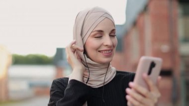 Cep telefonu tutan ve kulaklıkla müzik dinleyen modern Arap kadının portresi. Şehir caddesinde yürüyen akıllı telefon ve kulaklıklı Müslüman bir kadın. Kaldırımda tesettürlü güzel kız
