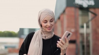 Müzik yayın platformu konsepti. Kablosuz kulaklık takan neşeli Müslüman kız, akıllı telefondan favori şarkıları dinliyor, kahve molası sırasında dans ediyor ve dinleniyor, şehirde yürüyor..
