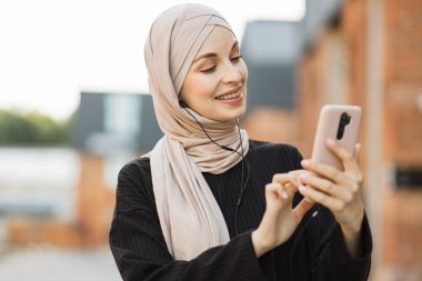 Cep telefonu tutan ve kulaklıkla müzik dinleyen modern Arap kadının portresi. Şehir caddesinde yürüyen akıllı telefon ve kulaklıklı Müslüman bir kadın. Kaldırımda tesettürlü güzel kız