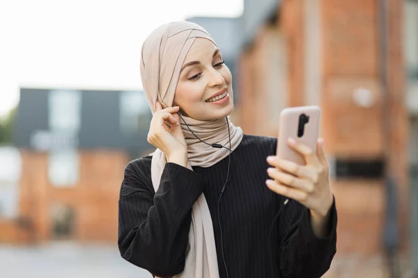 若いイスラム教徒の女性の音楽を聞きながら 街の通りを歩いてください ヘッドフォンをして携帯電話を手にしている ヒジャーブやカジュアルな服を着た — ストック写真