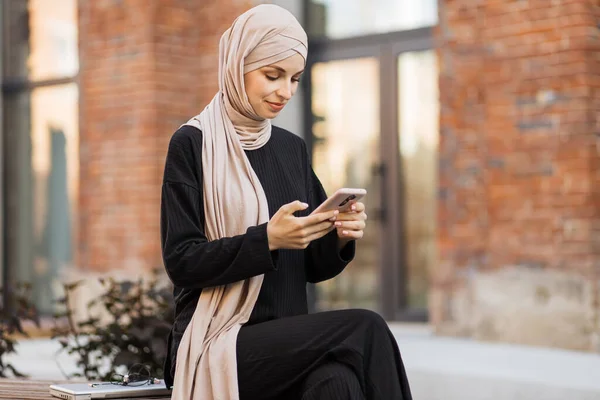 ヒジャーブ州のかなり笑顔のイスラム教徒のビジネス女性がベンチに座って ビデオチャット中にスマートフォンを使用しています 幸せなアラビア語の女の子座って屋外近く彼女の仕事のオフィスとチェック電子メール上のデジタルガジェット — ストック写真