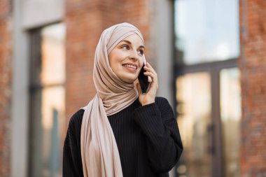 Siyah takım elbiseli, gülümseyen Müslüman kadın şehir caddesinde yürüyor ve telefonunu kullanıyor. Yazın tesettürlü güzel Arap kadın elinde cep telefonuyla sokakta yürüyor..