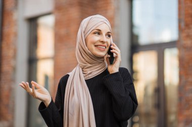Siyah takım elbiseli, gülümseyen Müslüman kadın şehir caddesinde yürüyor ve telefonunu kullanıyor. Yazın tesettürlü güzel Arap kadın elinde cep telefonuyla sokakta yürüyor..