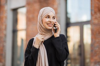 Siyah takım elbiseli, gülümseyen Müslüman kadın şehir caddesinde yürüyor ve telefonunu kullanıyor. Yazın tesettürlü güzel Arap kadın elinde cep telefonuyla sokakta yürüyor..
