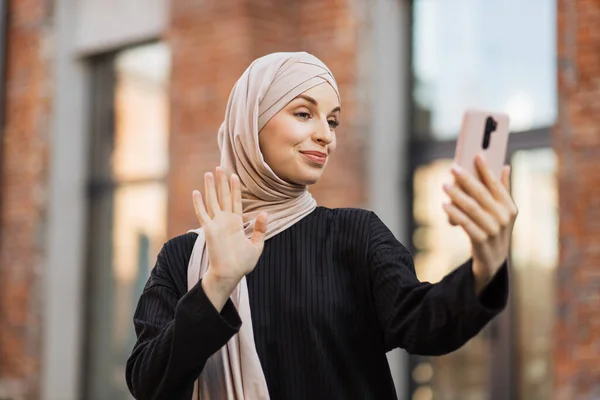 若い笑顔の幸せなイスラム教徒の女性が市内のスマートフォンを使ってビデオ通話をしています アラビア人女性ブロガー身に着けていますヒジャーブ語スマートフォンで話す彼女の聴衆と一緒に通りを歩く — ストック写真