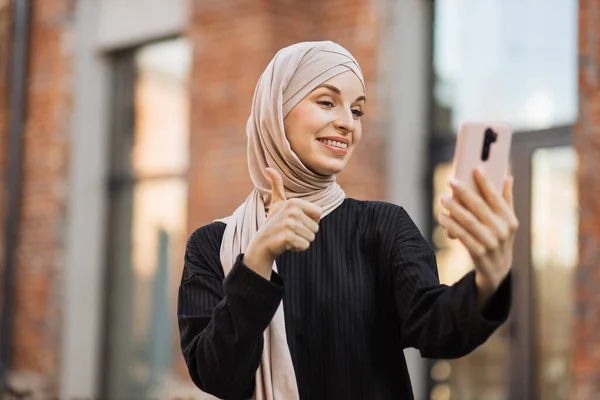 若い笑顔の幸せなイスラム教徒の女性が市内のスマートフォンを使ってビデオ通話をしています アラビア語の女性ブロガー身に着けていますヒジャーブ語スマートフォンで話すと彼女の聴衆と一緒に通りを歩いて親指を示す — ストック写真