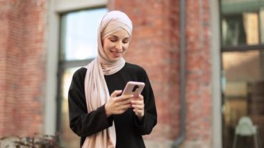 Güzel gülümseyen Müslüman iş kadını bankta oturuyor ve görüntülü konuşma sırasında akıllı telefon kullanıyor. Mutlu Arap kız iş yerinin yanında oturmuş dijital cihazla e-postalarını kontrol ediyor..