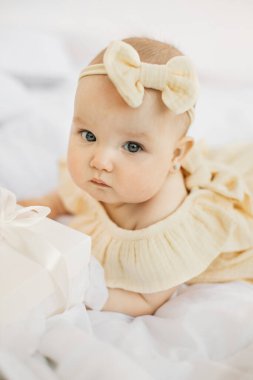 Sarı elbiseli mutlu küçük bir kız çocuğu ve parlak pencerenin yanındaki beyaz yatak odasındaki yatağın üzerinde başını eğiyor. Beyaz yatağa uzanmış, gülümseyen tatlı bir kız bebek. Bebek ürünleri konsepti.