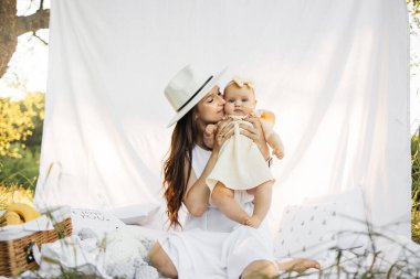 Genç anne kızıyla birlikte parkta ellerini tutarken yanaklarına dokunurken ve perdeli beyaz bir battaniyeye otururken dinleniyor. Aile pikniği. Anne ve küçük tatlı kızı yazın dinleniyor..