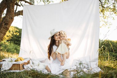 Beyaz şapkalı, siyah saçlı, beyaz şapkalı beyaz bir anne beyaz battaniye üzerinde kızıyla oturuyor. İki kişilik aile, yaz kampının tadını çıkarıyor..