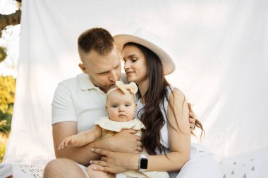 Neşeli genç ebeveynler beyaz battaniyenin üzerinde otururken dışarıda sevimli kızlarıyla oynuyorlar. Kızı öperken kucağında tutan adam. Boşluğu kopyala.