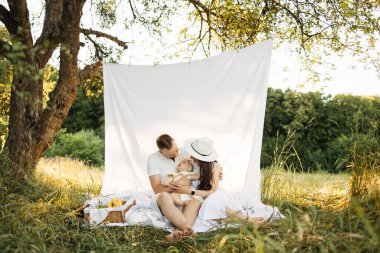 Portrait of young parents in white wear family look playing with six months old daughter on nature holding her in hands. Concept of family time and outdoors activity. Copy space.
