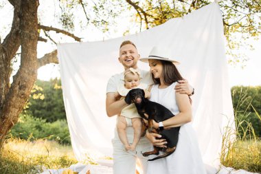 Beyaz elbiseli neşeli genç ebeveynler güzel küçük kız ve köpekle oynuyorlar beyaz battaniyenin üzerinde oturuyorlar resimli doğanın arasında. Doğayla iç içe geçen yaz zamanı. Aile kavramı.