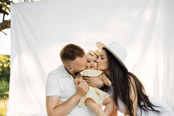 Young caucasian parents kissing pretty little daughter on cheeks from both sides, sitting together in background of white blanket. Happy family spending leisure time among green nature. Copy space.