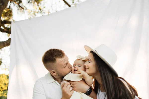 Young caucasian parents kissing pretty little daughter on cheeks from both sides, sitting together in background of white blanket. Happy family spending leisure time among green nature. Copy space.
