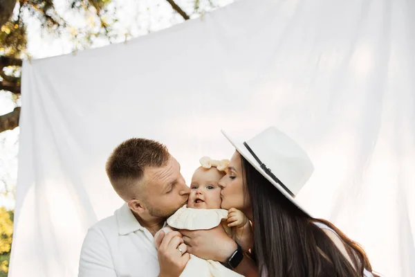 Young caucasian parents kissing pretty little daughter on cheeks from both sides, sitting together in background of white blanket. Happy family spending leisure time among green nature. Copy space.