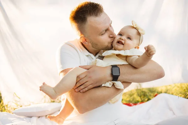 Portrait of happy father in white t-shirt kissing cheek of little daughter on background of white blanket during picnic on sunny summer day, close up view.