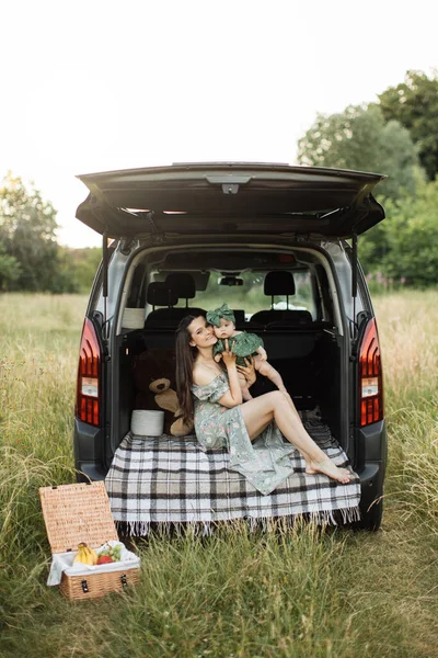 Cheerful young woman in green summer dress lifting up six month old baby girl while playing sitting on car trunk outdoors. Mother with daughter enjoying time spending together on fresh air.