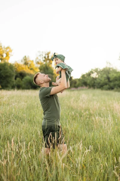 Attractive young father with green t-shirt and shorts lifting up his cute little daughter on hands playing together walking on meadows. Family time outdoors.