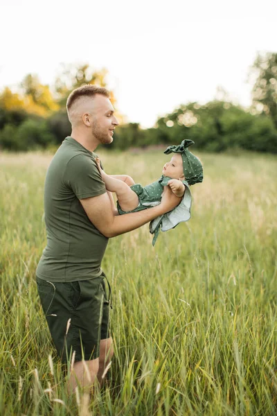Young man in green t-shirt and shorts lifting up his six month old baby girl while playing walking on field. Father with daughter enjoying time spending together on fresh air.