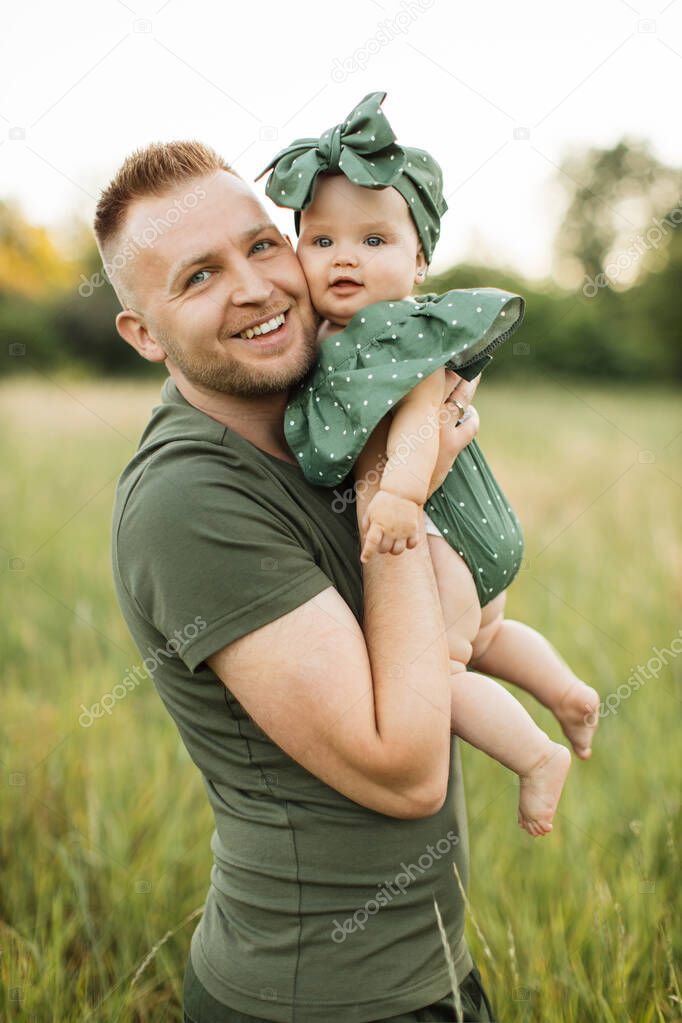 Portrait of young man smiling and looking at camera while walking in the meadow during picnic with his daughter on hands. Father enjoying every moment spending with his loving child.