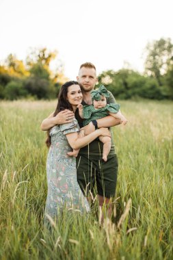 Portrait of happy young parents embracing little cute daughter while standing on the field on fresh air. Beautiful caucasian family having summer picnic on nature.