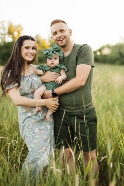 Joyful young parents playing with cute daughter outdoors while standing on background of green meadow. Man and woman holding baby girl on hands. Copy space.