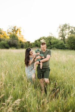 Beautiful and happy caucasian parents playing with their six months old daughter walking on green field. Positive family of three enjoying leisure time on fresh air.