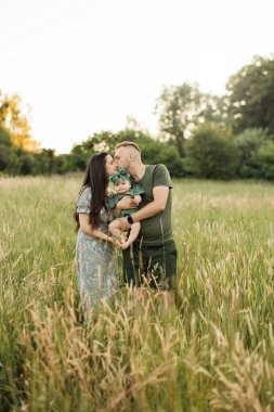 Neşeli, genç ebeveynler dışarıda duran şirin kızını kucaklıyor. Üç kişilik beyaz bir aile, gülümseyen bir baba annesini öpüyor ve yaz doğasında küçük bir kızı kucağına alıyor..