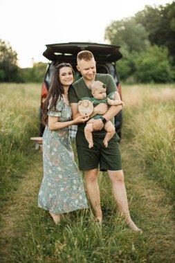 Güzel beyaz erkek ve kadın, arabalarının arka planında şirin küçük kızlarıyla birlikte güzel bir yaz otlağında dururlar. Temiz havada aile zamanı..