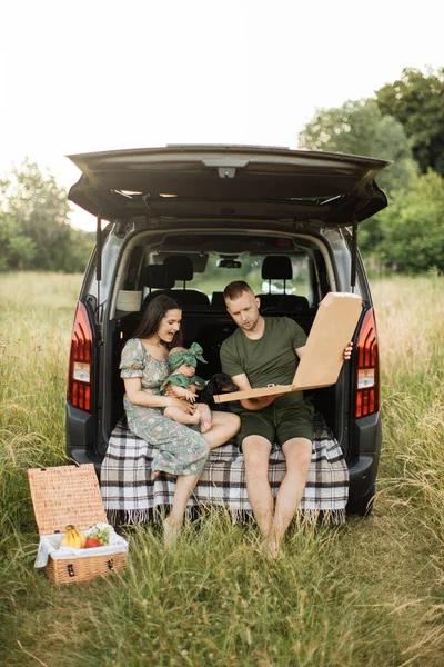 Happy young parents with dog and little daughter unpacking tasty pizza sitting on car trunk outdoors on the green field. Family of three spending weekend on nature.