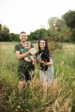 Beautiful and happy caucasian parents playing with their six months old daughter and dog walking on green field. Positive family of three enjoying leisure time on fresh air.
