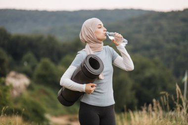 Yeşil Park 'ta dikilirken, tesettürlü ve aktif giyimli güzel bir bayanın şişe suyundan içişi. Müslüman kadın dışarıda yoga yaptıktan sonra dinleniyor ve ferahlatıyor..