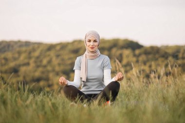 Huzurlu genç bayan tesettürde meditasyon yapıyor. Parktaki kameraya bakıyor. Yoga pozisyonunda mindere oturmuş güzel Müslüman kadın iç dengesini buluyor..