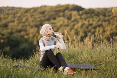 Attractive muslim woman in hijab resting while sitting on the mat and drinking water from a bottle outdoors in park . Healthy lifestyle concept.