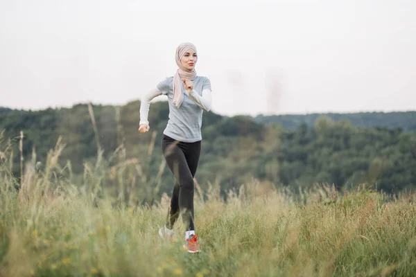 Happy arabian woman in hijab in activewear running at summer park. Young woman spending free time activity on fresh air. Smiling muslim female jogging during outdoors workout.